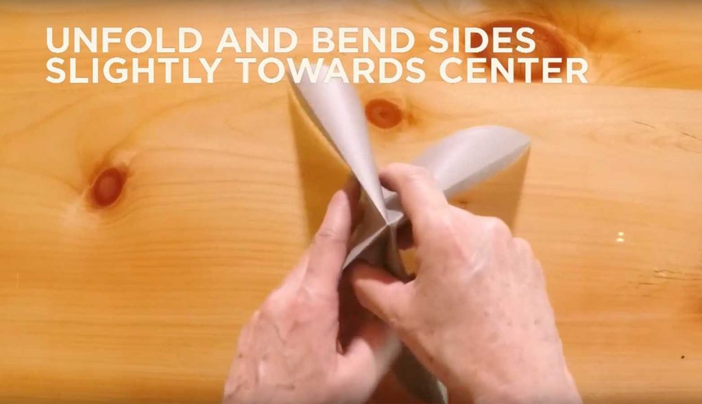 Paper folded into fourths with text saying, "Unfold and bend sides slightly towards center."