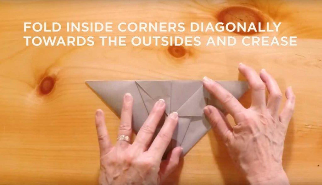 Paper being folded with text saying, "Fold inside corners diagonally towards the outsides and crease."