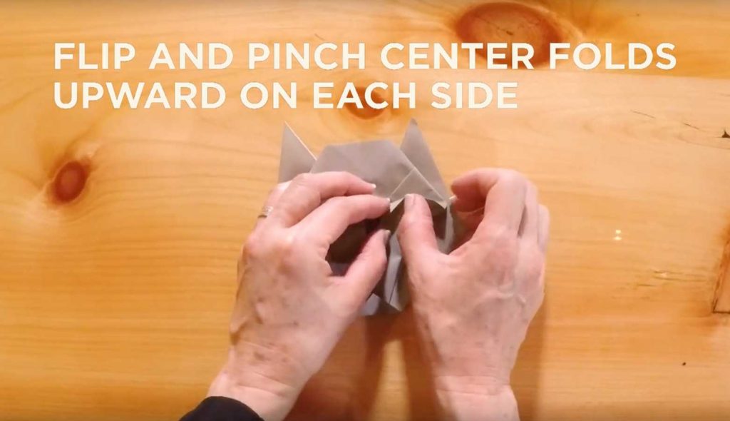 Paper being folded with text saying, "Flip and pinch center folds upward on each side."