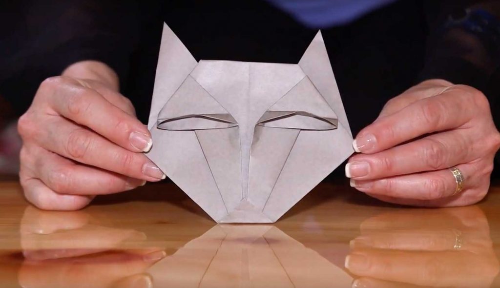 Completed origami animal wolf face displayed between a pair of hands holding it on a table.