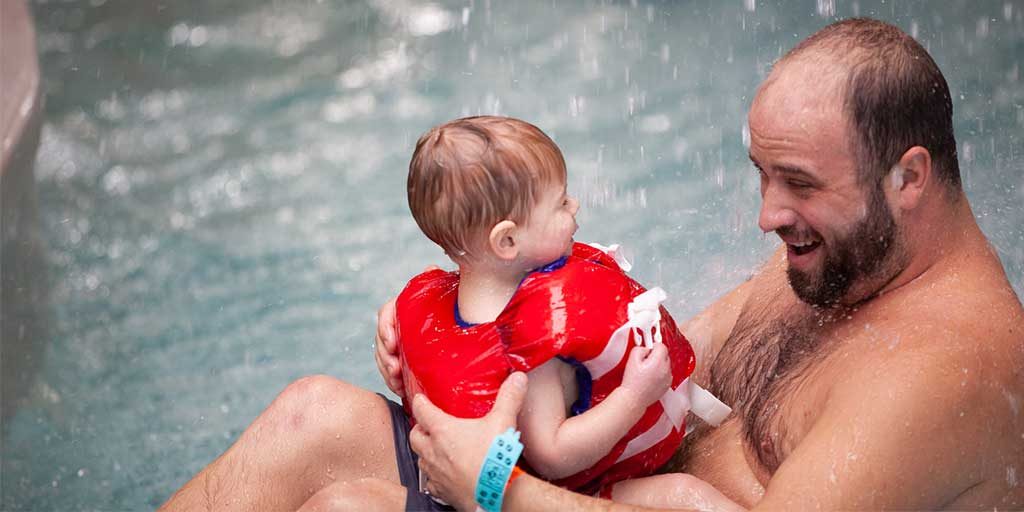 Dad smiling at little boy in a life jacket inside a pool