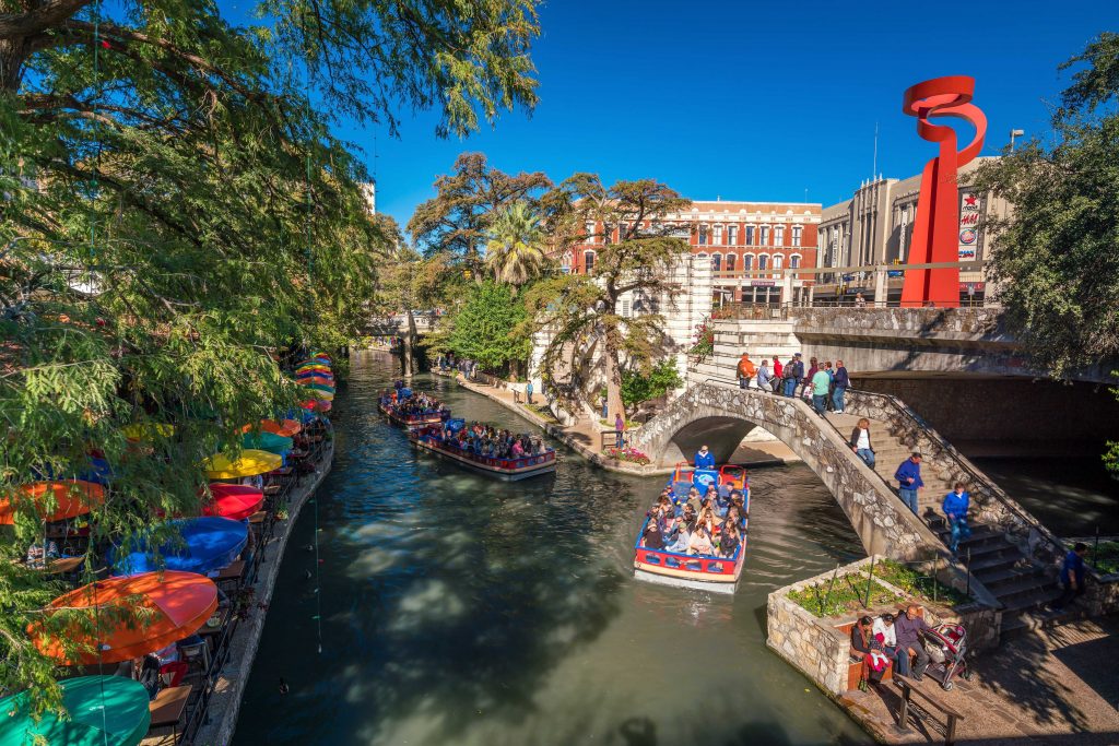 River walk in san antonio with boat with people on board going under river walk bridge.